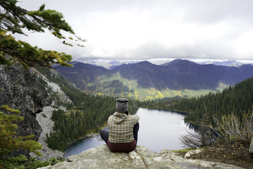woman sitting on ledge overlooking lake, mountains, and trees